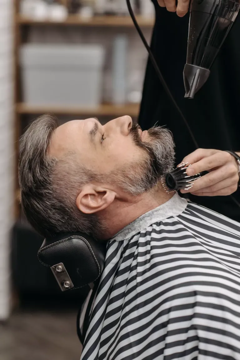 Hombre peinando la barba a otro hombre que está recostado en un sofá de barbería