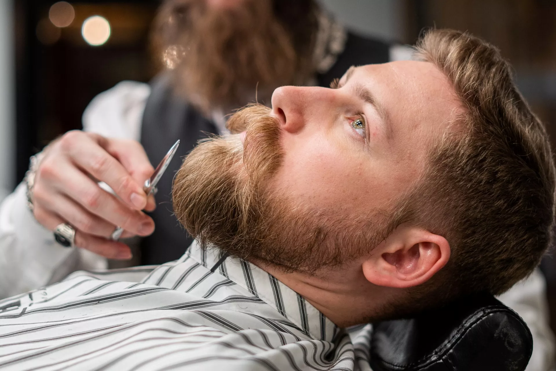 Hombre cortando la barba a otro hombre que está recostado en un sofá de barbería